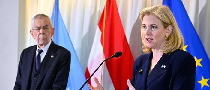Beate Meinl-Reisinger spricht vor einer Österreich- und einer EU-Flagge in ein Mikro, Alexander Van der Bellen sieht ihr zu.