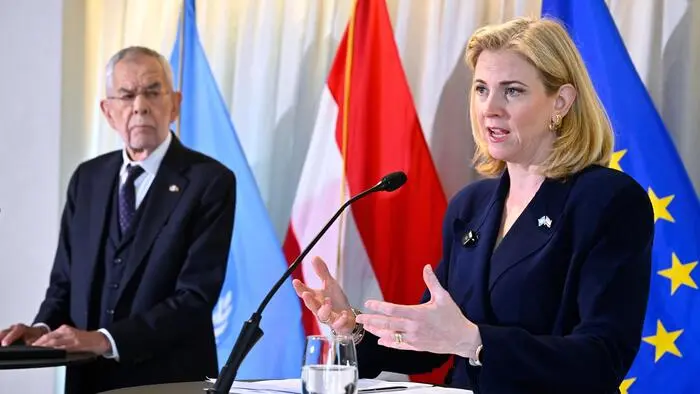 Beate Meinl-Reisinger spricht vor einer Österreich- und einer EU-Flagge in ein Mikro, Alexander Van der Bellen sieht ihr zu.