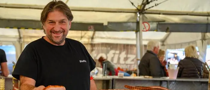 Ein Mann lacht in die Kamera und hält eine Bratwurst in der Hand