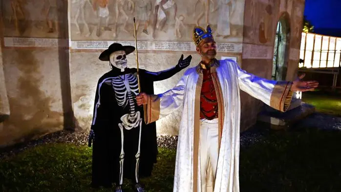 Vor dem Karner in Metnitz stehen zwei Darsteller: Einer ist als Tod verkleidet mit schwarzem Umhang, Hut und Totenschädel, der andere trägt einen weißen Umhang und eine Krone und symbolisiert damit den König.