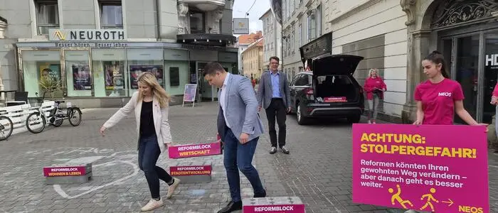 Ein Bild vom pinken Wahlkampfauftakt aus dem Mai 2026: Verena Garber (Listenzweite) und Alexis Pascuttini (Listendritter) tragen symbolisch einen schweren Stein mit der Aufschrift „Reformblock Wirtschaft“. Daneben ein großes pinkes Schild mit gelber Schrift und dem Titel „Achtung: Stolpergefahr!“