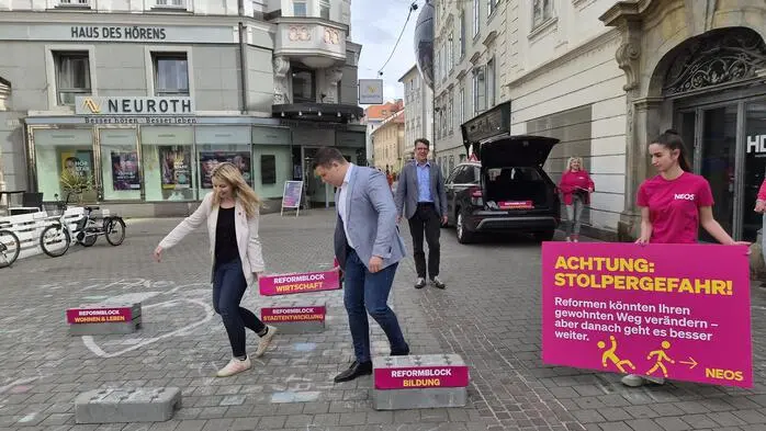 Ein Bild vom pinken Wahlkampfauftakt aus dem Mai 2026: Verena Garber (Listenzweite) und Alexis Pascuttini (Listendritter) tragen symbolisch einen schweren Stein mit der Aufschrift „Reformblock Wirtschaft“. Daneben ein großes pinkes Schild mit gelber Schrift und dem Titel „Achtung: Stolpergefahr!“