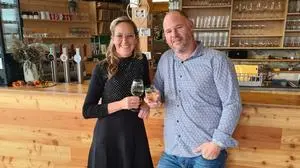 Eine Frau im schwarzen Kleid und ein Mann mit Hemd und blauer Jeans stehen mit einem Weinglas in der Hand vor einer Bar aus Holz | Petra und Bernhard Dobrounig