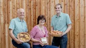 Zwei Männer und eine Frau mit Eiern, Nudeln und Mehl in der Hand lachen in die Kamera