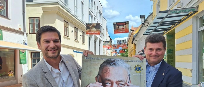 Zwei Männer halten ein Plakat, auf dem Kabarettist Andreas Vitasek zu sehen ist, in die Kamera 