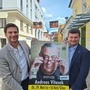 Zwei Männer halten ein Plakat, auf dem Kabarettist Andreas Vitasek zu sehen ist, in die Kamera 