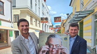 Zwei Männer halten ein Plakat, auf dem Kabarettist Andreas Vitasek zu sehen ist, in die Kamera 