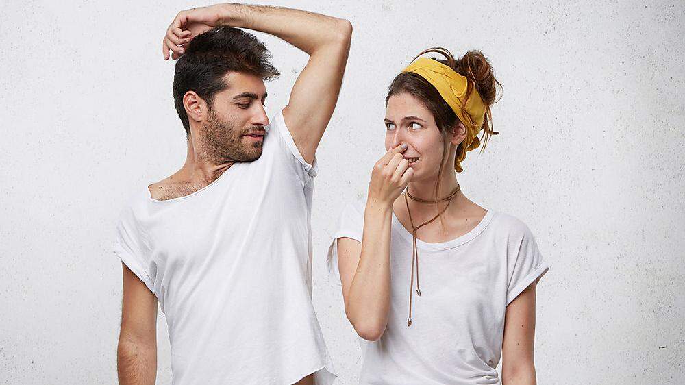 Female having disgusted look covering her nose with hand while smelling her husband`s armpit. Bearded man having nasty smell from sweat after running showing his wet underarm. Unpleasant smell