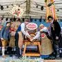 Viele Menschen in Tracht auf einer Bühne. Zwei stechen ein Bierfass an