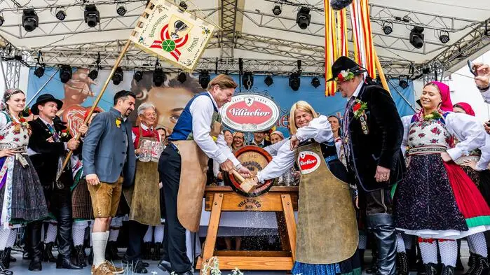 Viele Menschen in Tracht auf einer Bühne. Zwei stechen ein Bierfass an