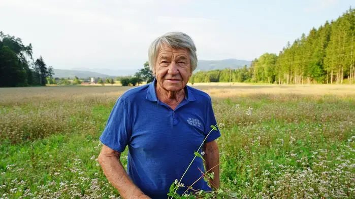 Ein Mann mit grauen Haaren steht auf einer Wiese und lacht in die Kamera