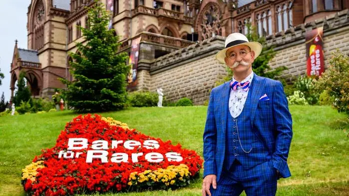 Horst Lichter steht in einem blaukarierten Anzug neben einem „Bares für Rares“-Schriftzug, der auf einem roten Blumenbeet angebracht ist.