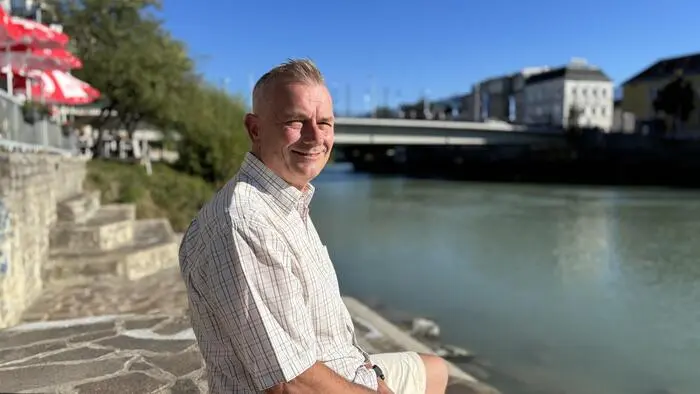 Ein Mann sitzt am Ufer eines Flusses, im Hintergrund sieht man eine Brücke
