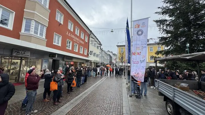 Eine Gruppe von Menschen steht entlang eines Platzes, in der Mitte wird eine Fahne gehisst | Am Dienstagvormittag wurde am Johannesplatz die Aktionsfahne zur Kampagne „Orange the World“ gehisst