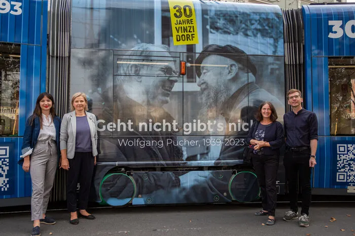Auf der Straßenbahn ist ein großes Bild vom kürzlich verstorbenen VinziDorf-Gründer Pfarrer Wolfgang Pucher mit einem Bewohner zu sehen. Darunter steht der Leitspruch Puchers: Geht nicht, gibt's nicht.