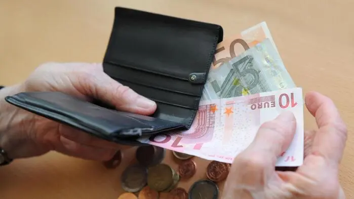 Die Hände einer alten Frau ziehen Geldscheine aus einer Brieftasche. Auf dem Tisch darunter liegen Münzen