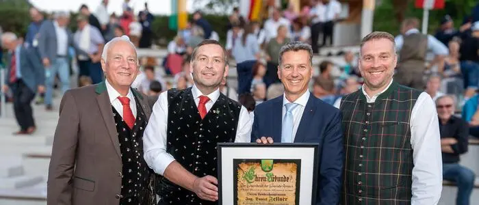 Vier Männer in Tracht stehen vor einer Tribüne und halten gemeinsam eine Urkunde in die Kamera