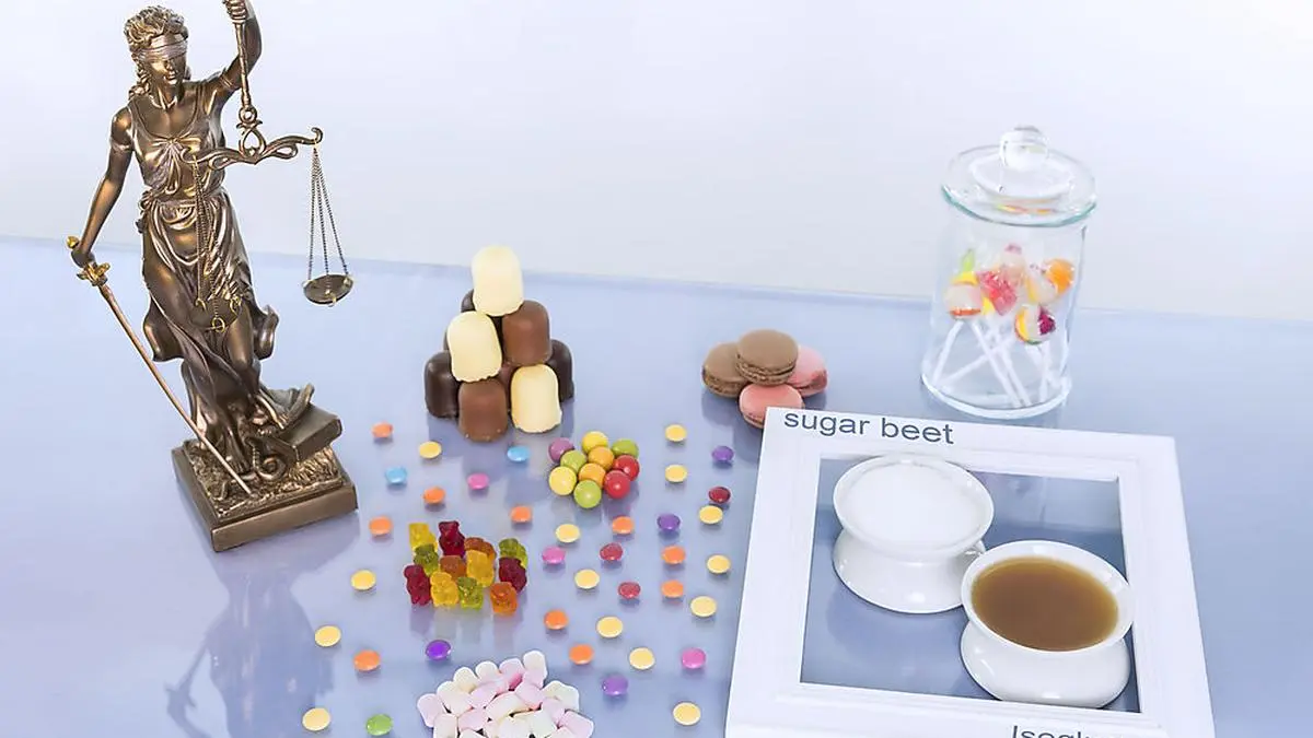 Justitia Statue steht auf dem Tisch mit vielen süßen Lebensmitteln. Zum Vergleich stehen dort  jeweils eine Schale mit Zucker und eine Schale mit Industriezucker nebeneinander. Konzeptbild