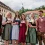 Vier Frauen und zwei Männer, alle in Tracht, lachen in die Kamera. Im Hintergrund ist der Grazer Uhrturm zu sehen.
