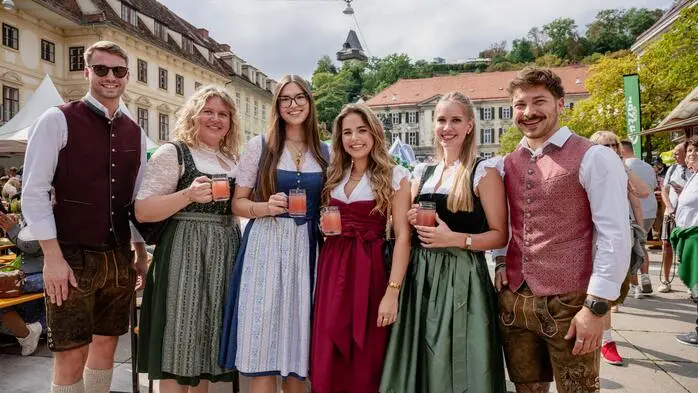 Vier Frauen und zwei Männer, alle in Tracht, lachen in die Kamera. Im Hintergrund ist der Grazer Uhrturm zu sehen.
