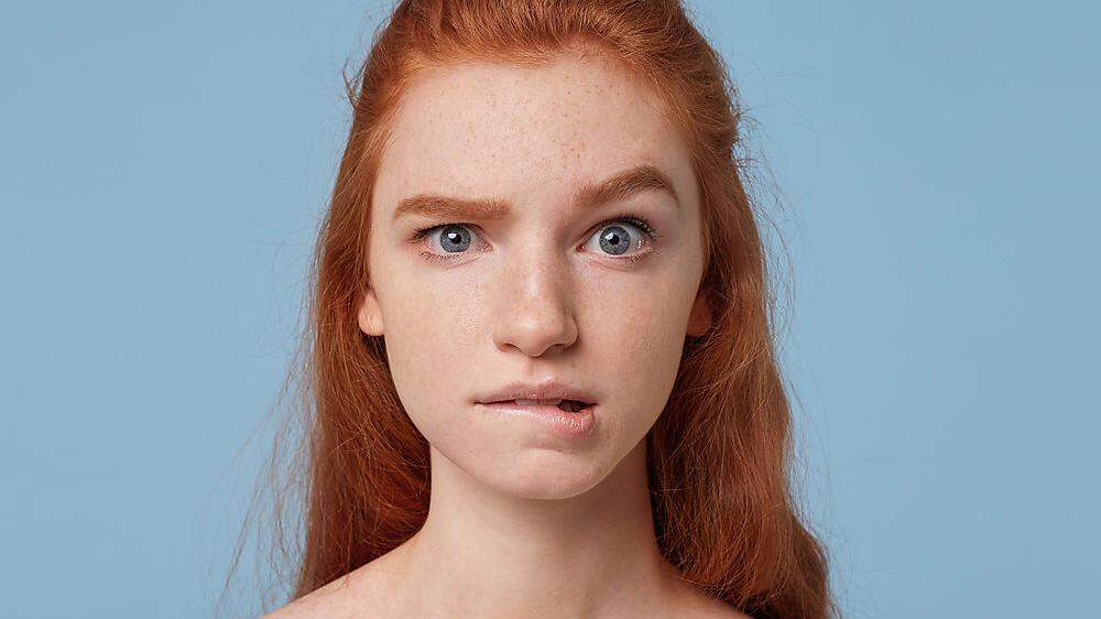 Young beautiful red-haired teen girl looks concerned puzzled in a panic, one eyebrow raised bit her lower lip isolated on a blue background.