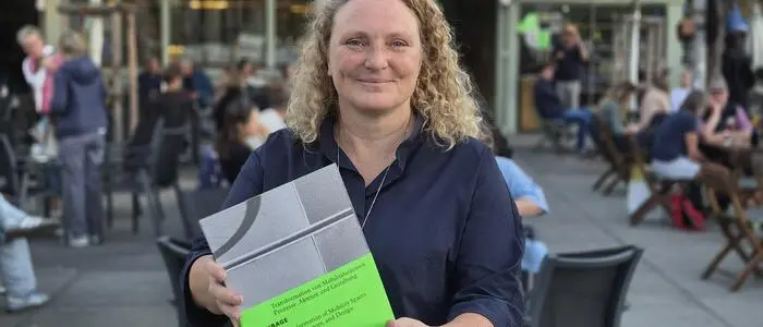 Frau mit blonden Locken steht am Marktplatz und hält ein Buch hoch