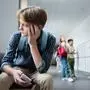 depressed boy with smartphone sitting alone in school corridor near teenagers on blurred background