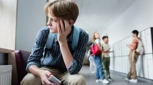 depressed boy with smartphone sitting alone in school corridor near teenagers on blurred background