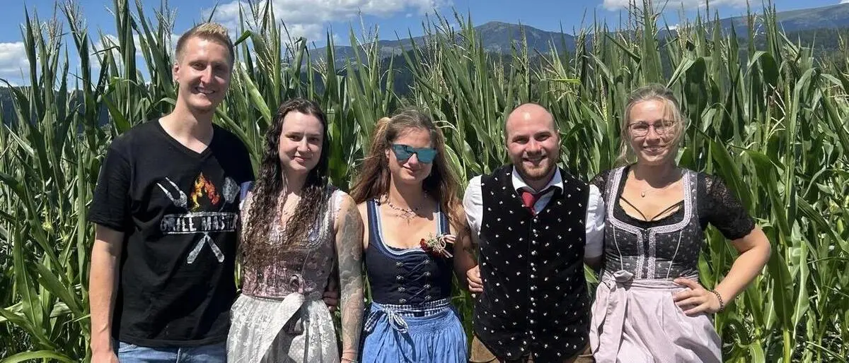 Drei Frauen und zwei Männer in Tracht stehen vor einem Maisfeld und lachen in die Kamera