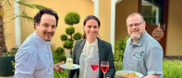 Das Wirtepaar Peter und Susanne Moser und Mitveranstalter Willie Wurzer stehen nebeneinander und halten Teller mit Essen und Getränken in den Händen
