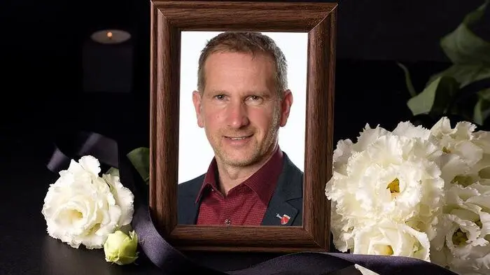 Portärt eines Mannes im Anzug in einem Bilderrahmen. Davor weiße Blumen und schwarze Schleifen
