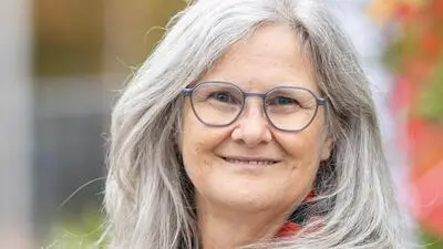 Eine Frau mit grauen langen Haaren und Brille lacht in die Kamera