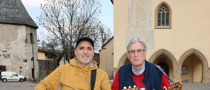Zwei Männer, einer mit Gitarre und der andere mit einer Ukulele in der Hand, stehen auf einem großen Platz, im Hintergrund ist eine Kirche
