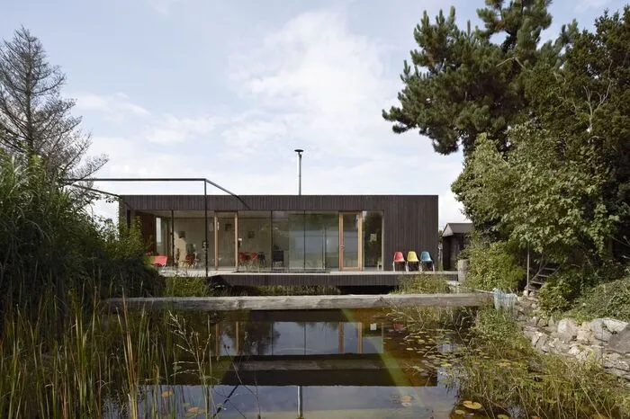 Das "beste Haus" in Oberösterreich ist das "Haus am Teich", geplant von "Hammerschmid, Pachl, Seebacher – Architekten". Die Jury versteht dieses  Haus als eine gelungene Antwort auf die Frage, wie wenig Kubatur man zum Leben braucht, und ein „vierfaches Paradebeispiel“ für nachhaltiges Bauen. "Erstens: Das große elterliche Grundstück wurde mit einem Wohnhaus neben dem bestehenden Schwimmteich nachverdichtet. Zweiten wurden dabei ökologische Baumaterialien und Dämmstoffe verwendet, und zwar - drittens - in minimalem Ausmaß. Viertens ist das Haus bei Bedarf mobil, Statik und Dimensionen ermöglichen den Abtransport und Aufbau an anderer Stelle. Durch die aufgeständerte Typologie schmiegt sich der Baukörper an die Topographie, ohne den bestehenden Geländeverlauf zu verändern, unter dem Wohnhaus gibt es Platz für PKW-Abstellplätze und Brennholz. Die Fassade aus sägerauhen Fichtenbrettern ist mit schwedischer Schlammfarbe gestrichen, im Innenraum sind Boden- Decken-Wand- und Möbeloberflächen mit weiß geöltem Birkensperrholz ausgeführt."