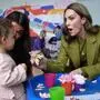 Das Bild zeigt eine lebhafte Szene in einem bunt gestalteten Raum, vermutlich einer Kindertagesstätte oder einem Gemeinschaftszentrum.
Im Vordergrund sieht man Prinzessin Kate in einem olivgrünen Blazer, die sich einem kleinen Kind zuwendet. Sie hält eine Blume in der Hand und schaut das Kind mit einem erstaunten, freundlichen Ausdruck an – ihr Gesicht zeigt Interesse und Begeisterung. Das Kind trägt einen rosafarbenen Strampler und hält ebenfalls etwas in der Hand. Eine weitere Frau, teils hinter dem Kind, beobachtet die Situation lächelnd. | „Obwohl digitale Geräte versprechen, uns miteinander zu verbinden, bewirken sie oft das Gegenteil“, schreibt Kate 