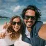 A pair of lovers take a selfie on a tropical beach. The guy and the girl cheerfully smile at the phone camera on the background of the sea. Newlyweds and tropical lagoon with green mountains and cloudy sky.