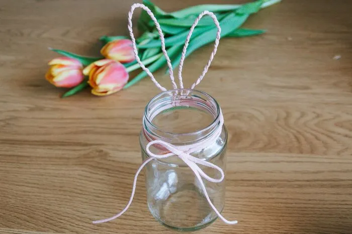 Die geformten Hasenohren am Einmachglas anlegen und fixieren, indem man das zweite Stück vom Band ein paar Mal um das Glas herumwickelt. Vorne mit einer Schleife fixieren.