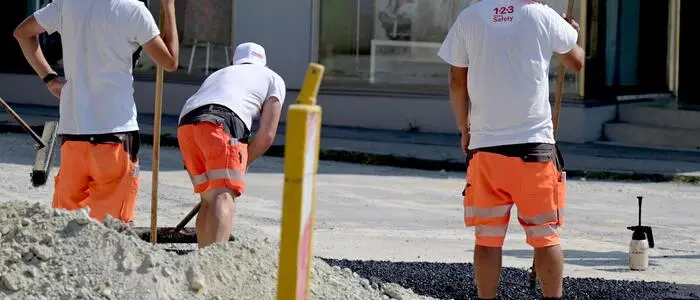 Man sieht drei Straßenarbeiter von hinten bei der Arbeit, zwei von ihnen wirken erschöpft