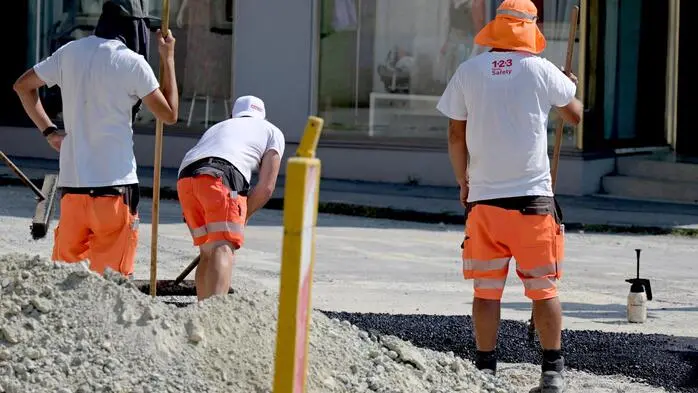 Man sieht drei Straßenarbeiter von hinten bei der Arbeit, zwei von ihnen wirken erschöpft
