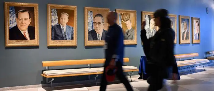 Besucher gehen an einer blauen Wand mit gerahmten Porträts früherer UN-Generalsekretäre vorbei. Die Gemälde hängen in einer Reihe über Sitzbänken; zwei Personen im Vordergrund sind durch ihre Bewegung leicht verschwommen.