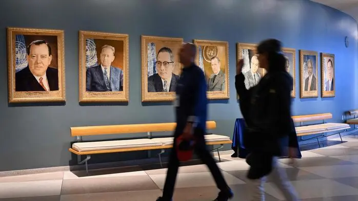Besucher gehen an einer blauen Wand mit gerahmten Porträts früherer UN-Generalsekretäre vorbei. Die Gemälde hängen in einer Reihe über Sitzbänken; zwei Personen im Vordergrund sind durch ihre Bewegung leicht verschwommen.