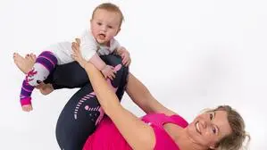 Eine blonde Frau im sportlichen Outfit hält ein Baby hoch und lacht in die Kamera