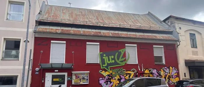 Außenansicht eines roten einstöckigen Gebäudes, davor stehen ein Moped und zwei Autos