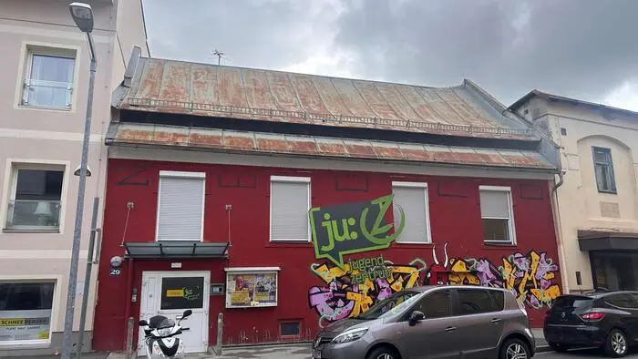 Außenansicht eines roten einstöckigen Gebäudes, davor stehen ein Moped und zwei Autos