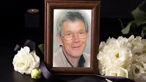 Ein Foto von einem Mann mit Brille in einem Bilderrahmen, daneben liegen weiße Blumen