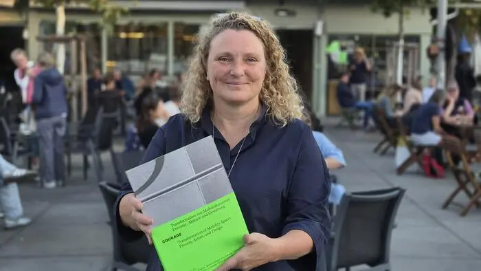 Frau mit blonden Locken steht am Marktplatz und hält ein Buch hoch
