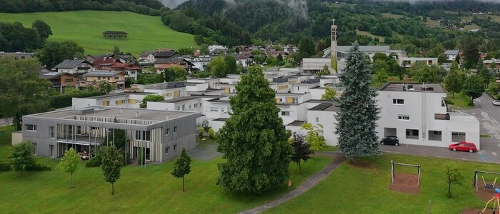 Man sieht weiße, graue und braune Gebäude aus der Luft fotografiert in einer wolkigen Landschaft mit Wiesen und Bäumen | Das SOS Kinderdorf in Nussdorf-Debant