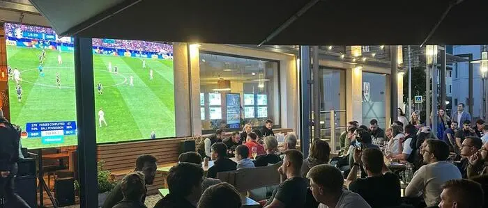 Viele Menschen sitzen und stehen auf einer Terrasse und schauen auf eine Leinwand, dort ist ein Fußballspiel zu sehen