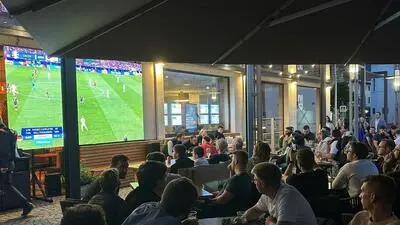 Viele Menschen sitzen und stehen auf einer Terrasse und schauen auf eine Leinwand, dort ist ein Fußballspiel zu sehen