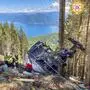 Das Bild zeigt die Folgen eines tragischen Unfalls mit der Seilbahn Stresa-Mottarone, der sich am 23. Mai 2021 ereignete. Im Zentrum des Bildes liegt das Wrack der Seilbahnkabine auf einem bewaldeten Berghang. Sie ist schwer beschädigt und liegt auf der Seite. Mehrere Rettungskräfte in leuchtend gelben und grünen Uniformen sind an der Unfallstelle zu sehen. Sie scheinen die Umgebung zu untersuchen und an der Bergung beteiligt zu sein. Der Berghang ist dicht bewaldet, mit hohen Nadelbäumen. Der Boden ist mit abgebrochenen Ästen und Trümmern bedeckt. Im Hintergrund des Bildes erstreckt sich eine malerische Landschaft mit einem See (Lago Maggiore) und einer Gebirgskette. Das sonnige Wetter und der klare Himmel bilden einen starken Kontrast zu der tragischen Szene. Am oberen rechten Bildrand ist ein rundes Emblem zu sehen, das das Zeichen des „Corpo Nazionale Soccorso Alpino e Speleologico“ (Italienische Bergrettung) zeigt. | Die abgestürzte Gondel am Berg Mottarone am Lago Maggiore